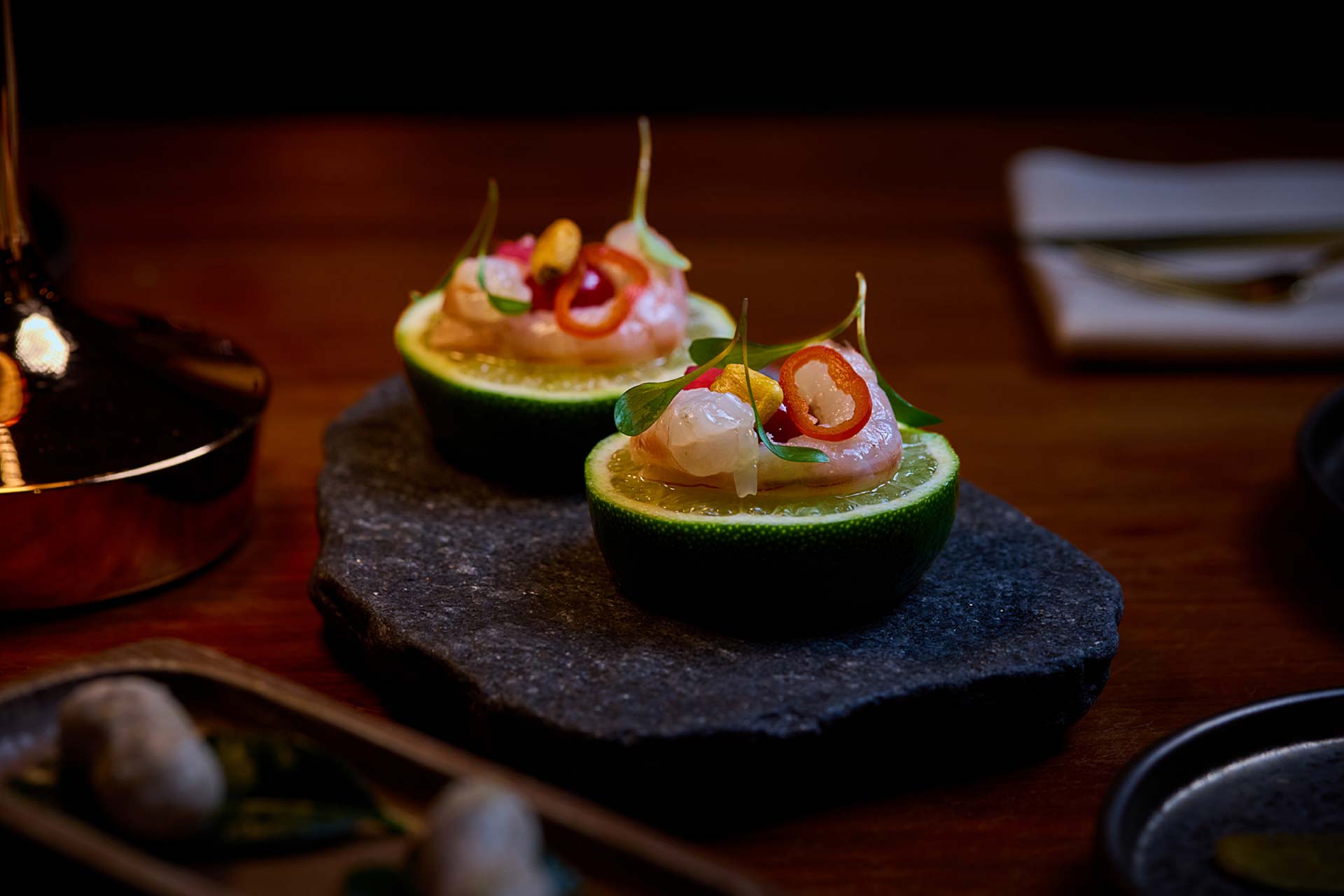 Two lime halves topped with raw seafood, citrus slices, peppers, and greens on a dark stone platter.