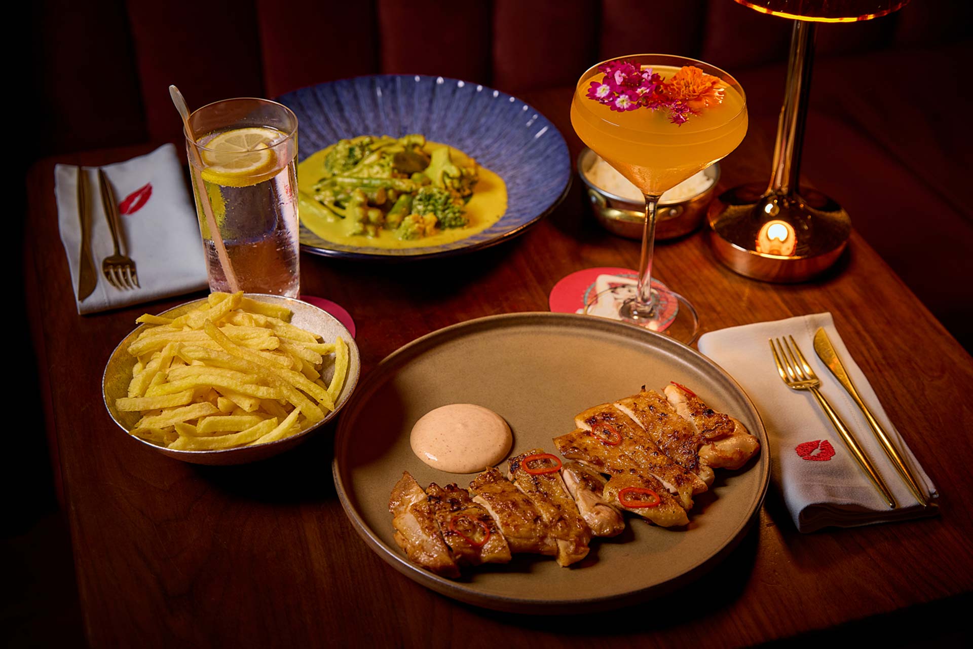 Table setting with grilled sliced chicken, fries, a glass of lemon water, and a yellow cocktail with flowers under warm lighting
