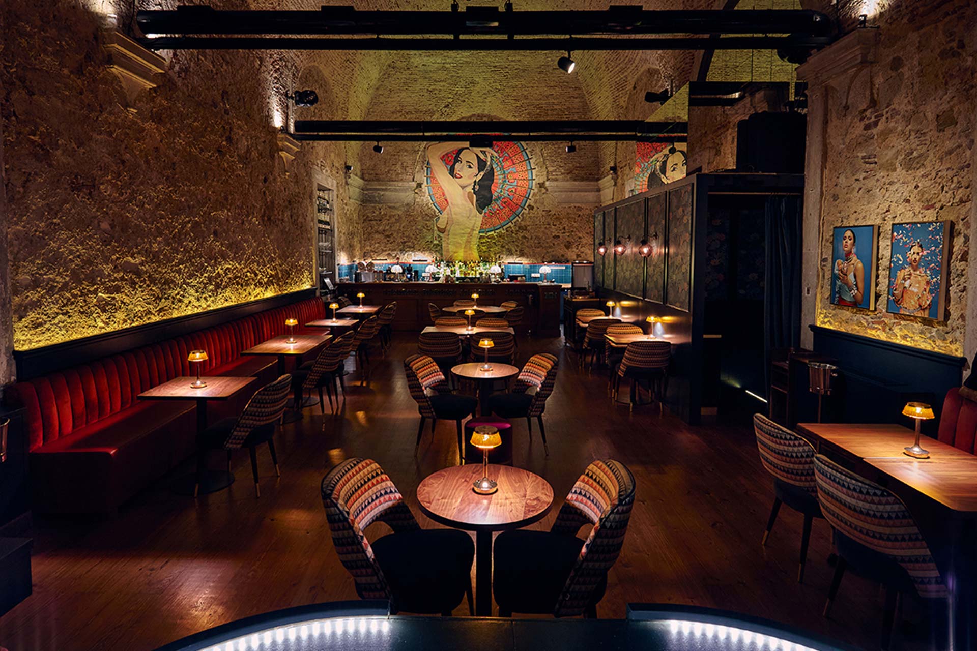 Cozy bar and dining area with exposed brick walls, warm lighting, round wooden tables, and striped chairs, centered by a large mural of a woman on the back wall.