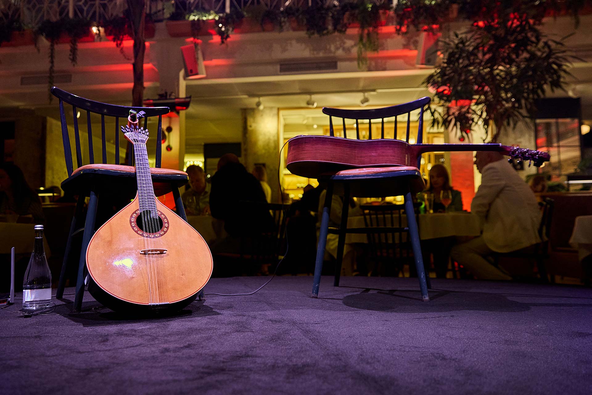 Lute on the floor leaning against a chair on a dimly lit stage, with a guitar resting on another chair behind it and a bottle nearby.