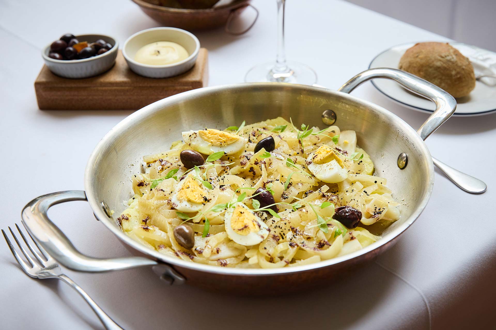 Pasta with sliced hard-boiled eggs, black olives, and fresh herbs served in a skillet on a white table.