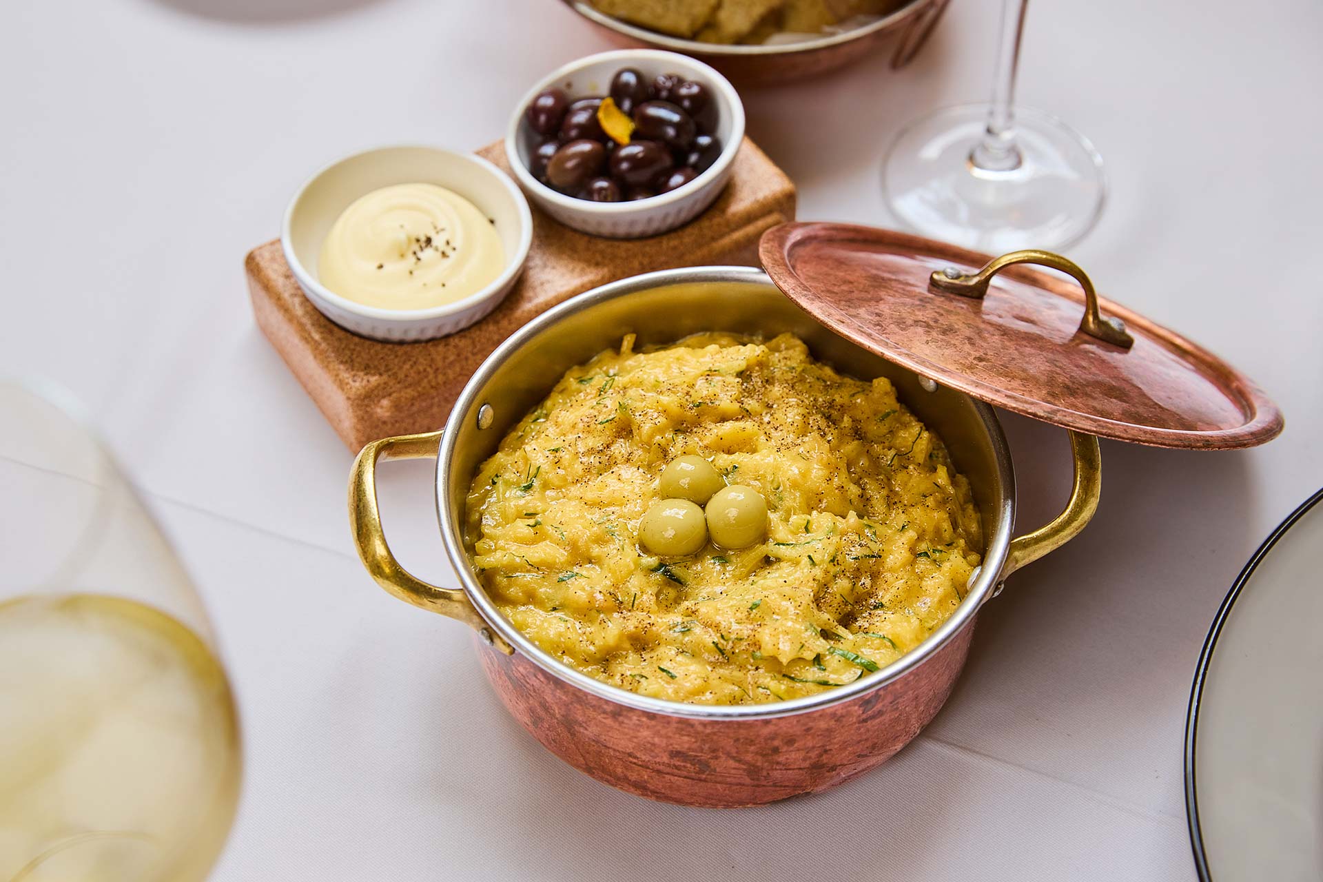 Copper pot of herb risotto with green olives, lid ajar, on a white table with side bowls of sauce and olives and a wine glass nearby