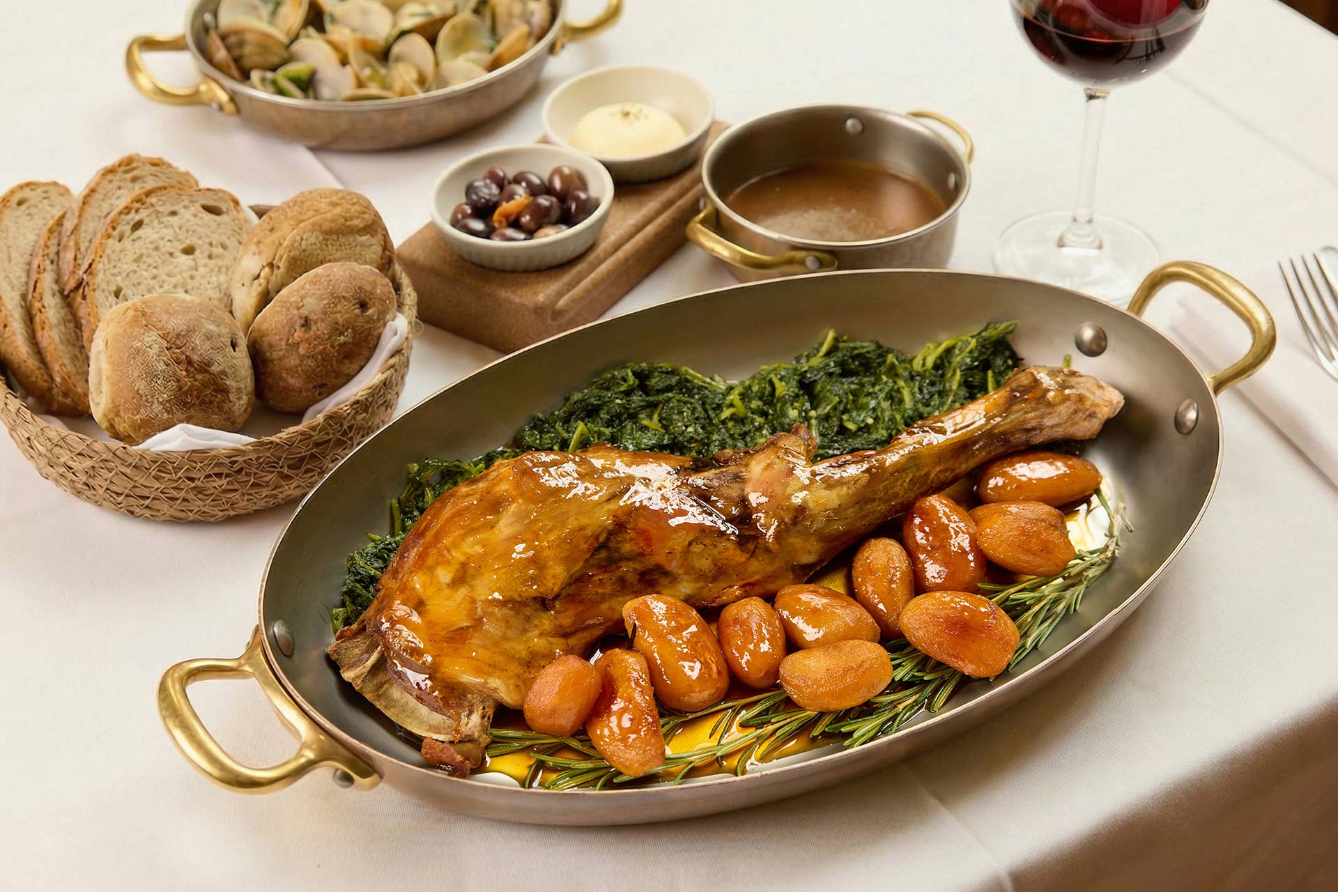 Roasted turkey leg with glazed potatoes and sautéed greens on a gold-rimmed platter, with bread basket and wine glass nearby.