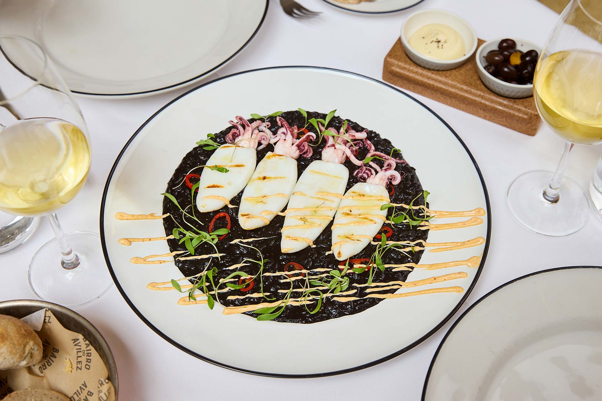 Plate of black squid ink risotto topped with grilled white fish slices, shrimp, and microgreens, drizzled with orange sauce and red pepper rings on a white table.