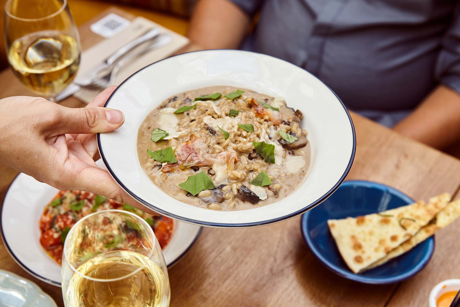 A hand holds a bowl of creamy mushroom risotto topped with fresh herbs as wine glasses and other dishes sit on a wooden table.