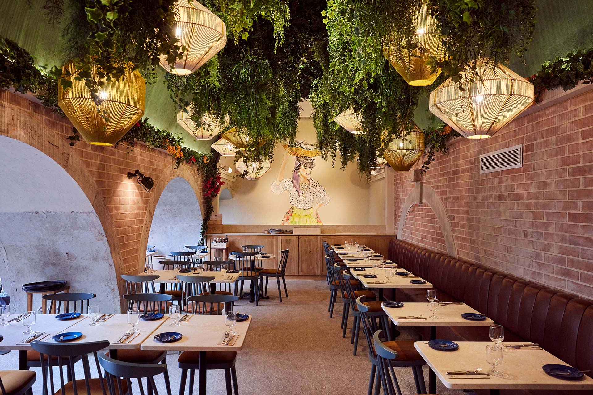 Restaurant dining room with brick arches, hanging greenery and warm wicker lanterns, and a mural of a flamenco dancer on the back wall.