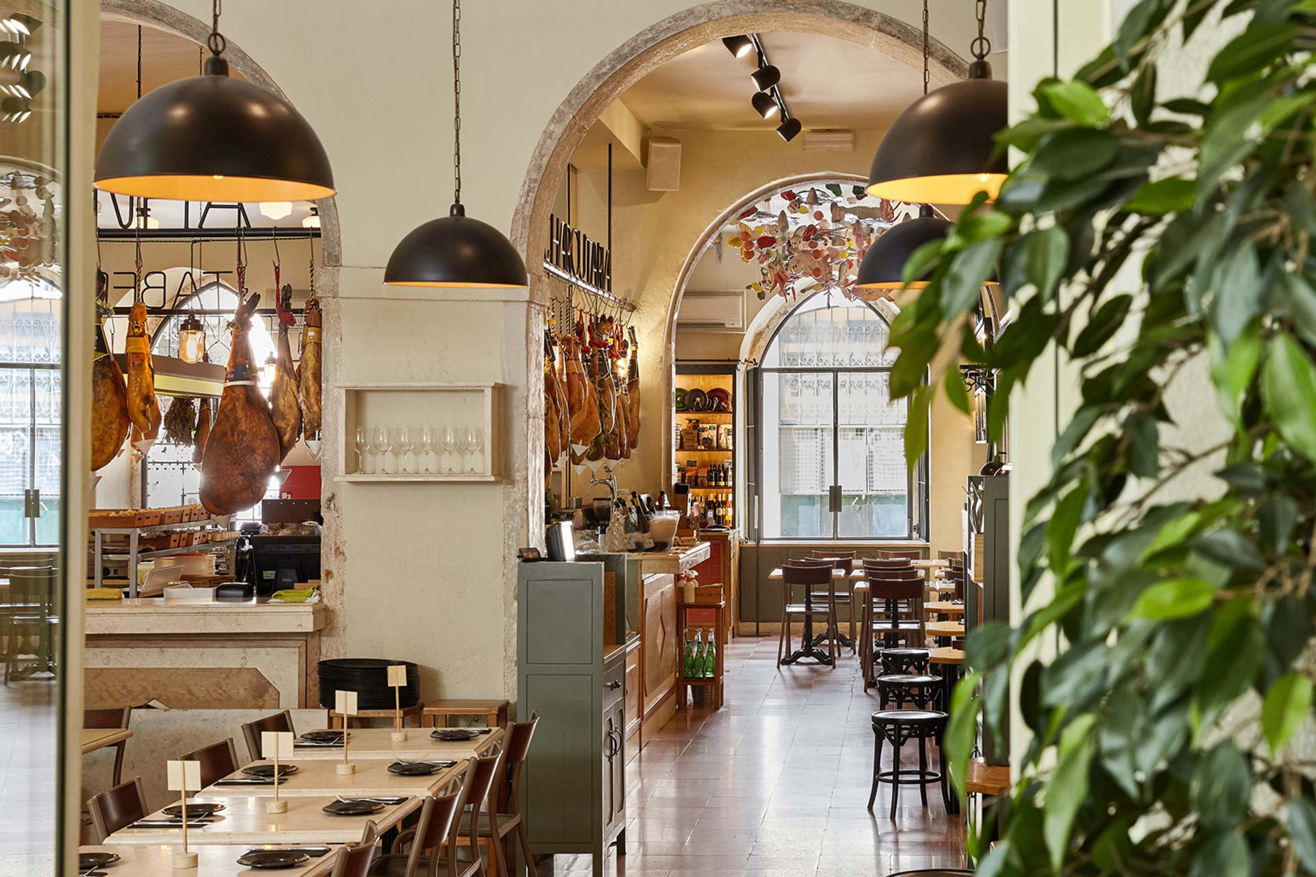 Dining room of a rustic restaurant with arches, hanging cured hams, and warm pendant lights; green plants frame the right side.