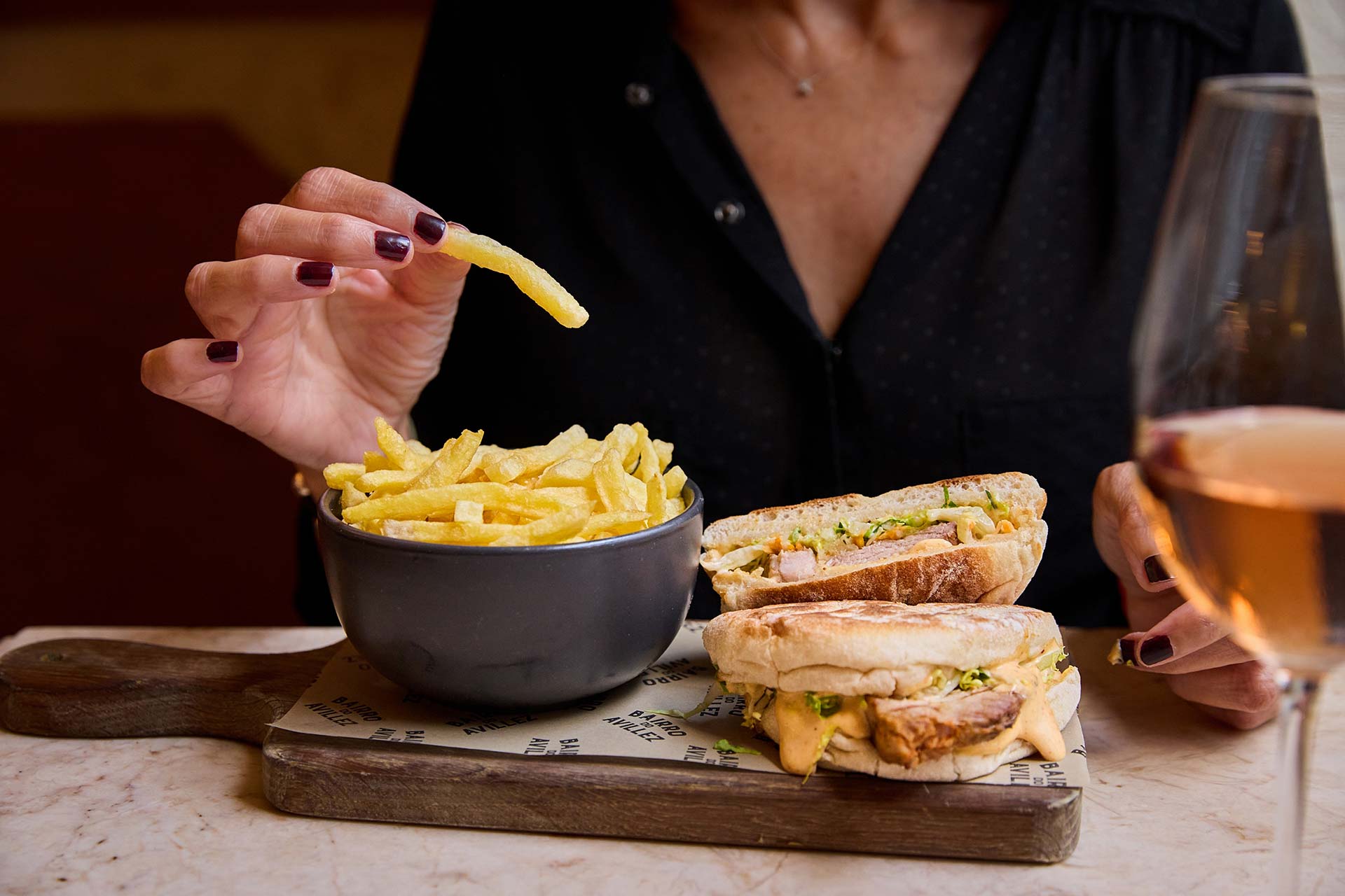 Hand picking up a French fry from a bowl, with a sandwich and wine glass nearby on a wooden board.