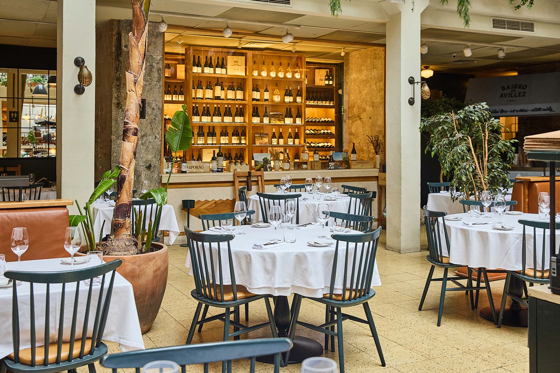Restaurant dining area with white-tablecloth tables, green chairs, and a backlit wine display wall behind glass shelves.