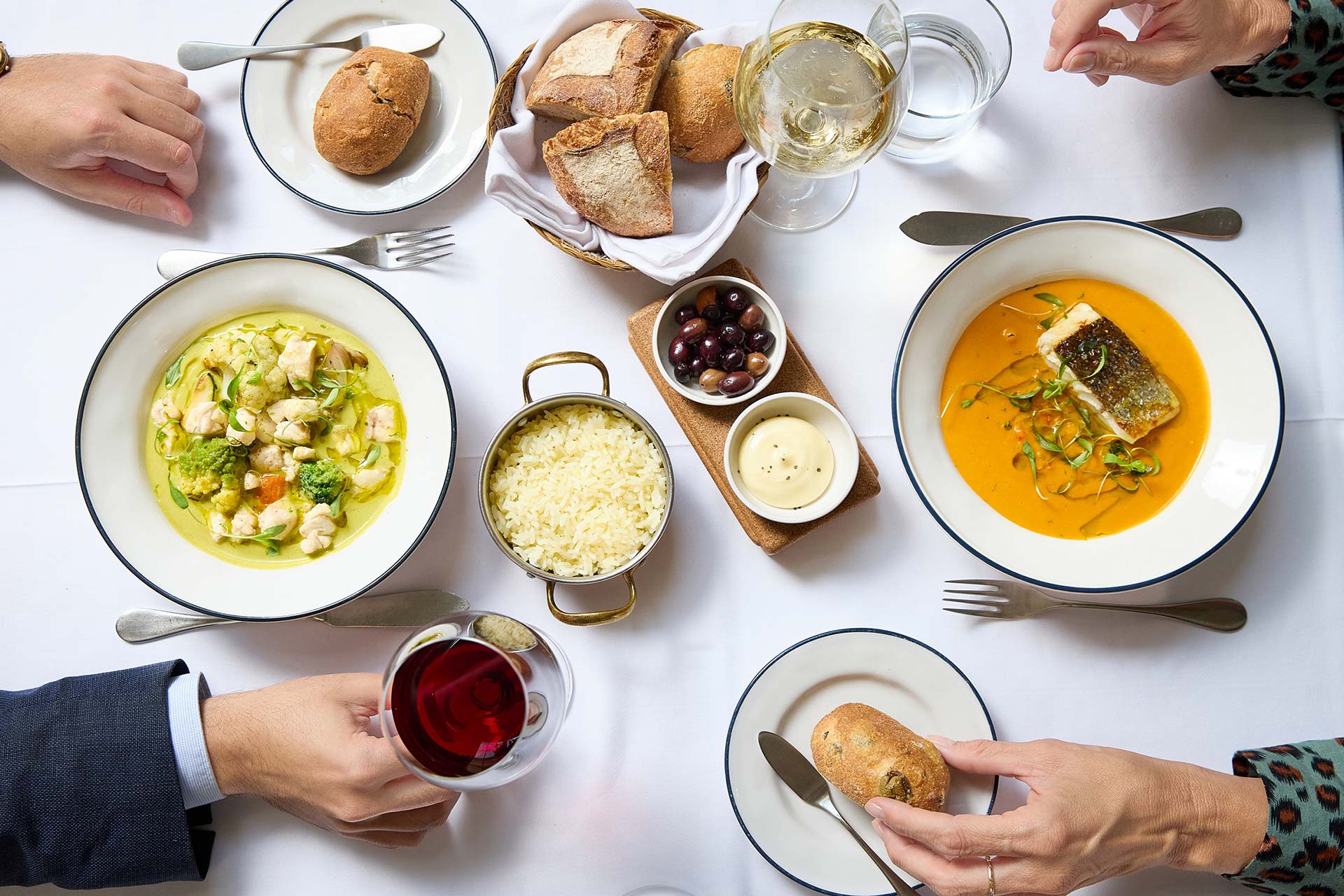 Top-down view of a dining table with two bowls of soup, bread basket, rice, olives, and a glass of white wine, with hands reaching for food.