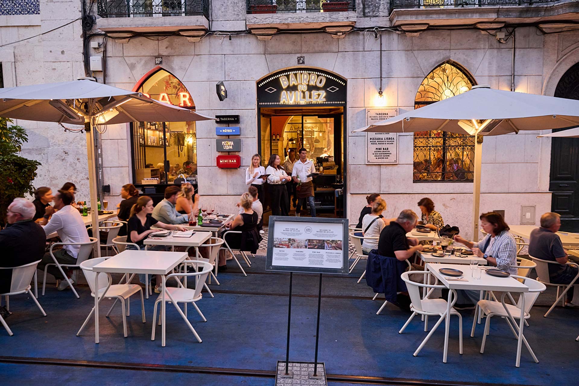 Outdoor restaurant terrace with diners at white tables under large umbrellas, in front of a stone building with a lit 'Bairro do Avillez' sign.