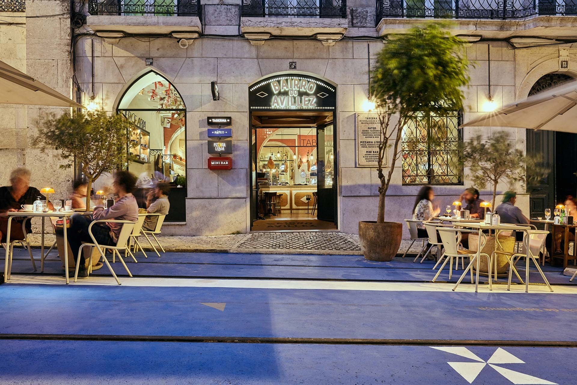 Outdoor cafe on a pedestrian street at dusk with people seated, blue pavement, and a central arched doorway labeled 'Bairro do Avillez'