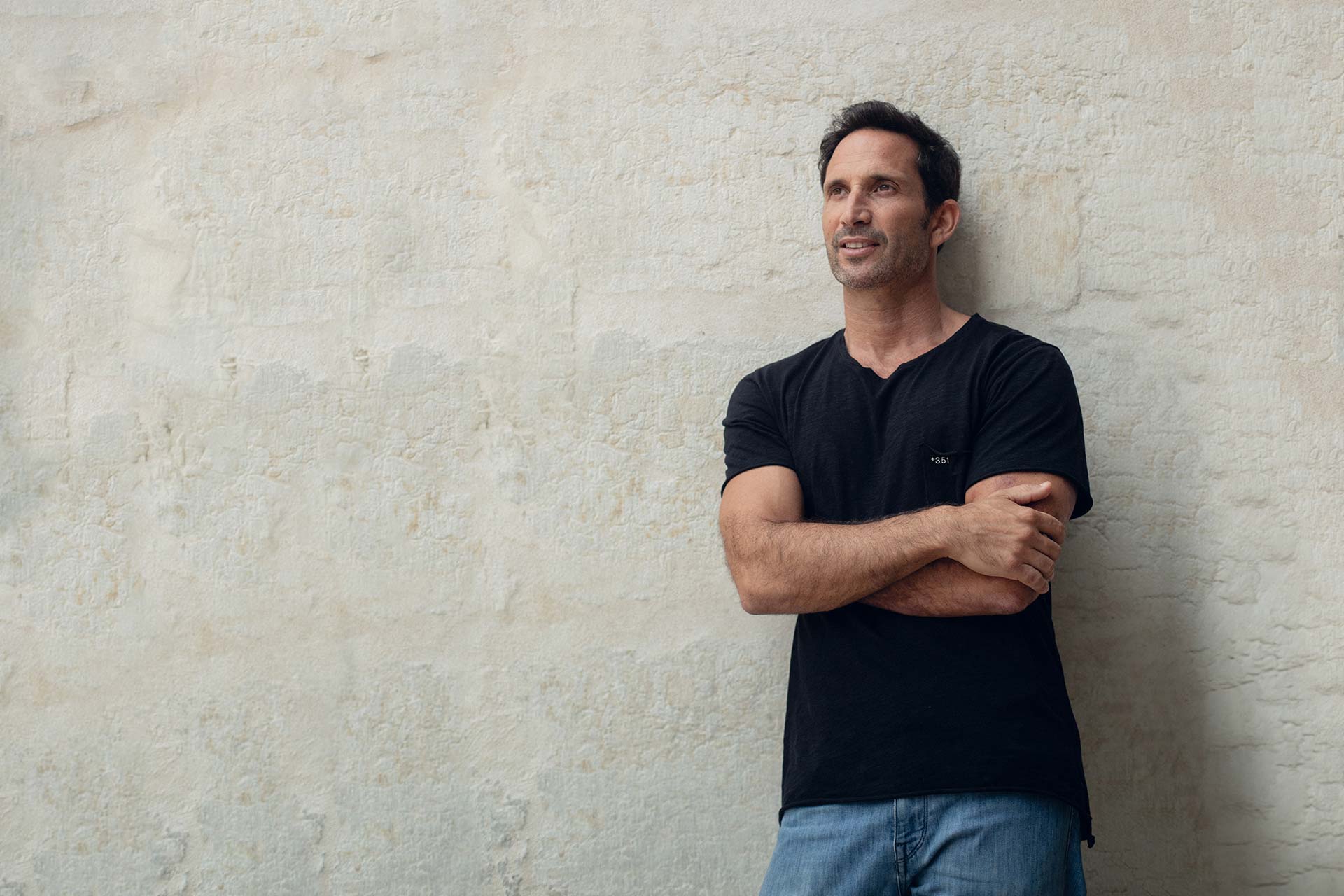 Man leaning against a light stone wall with arms crossed, wearing a black t-shirt and jeans