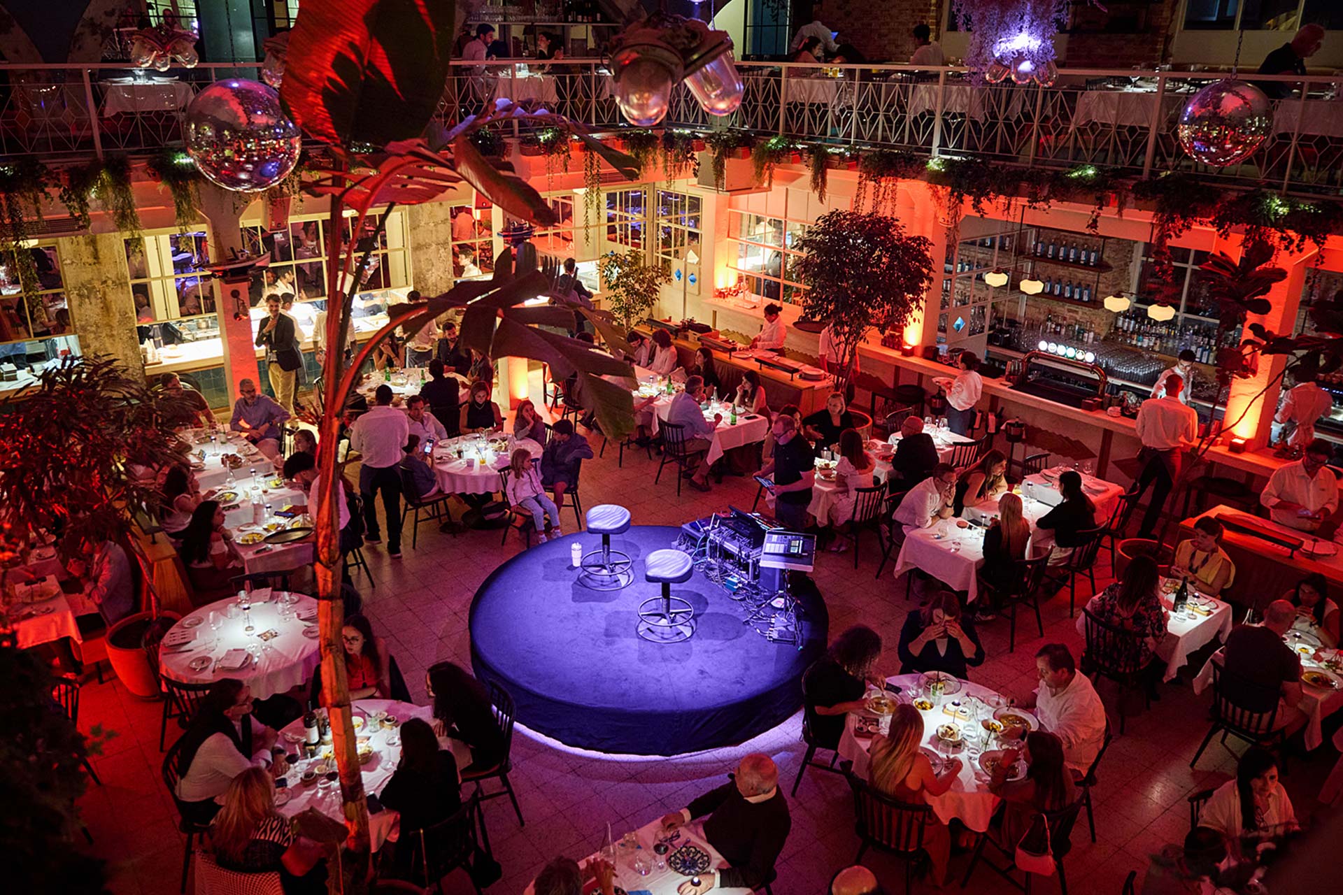 Busy restaurant interior viewed from above with many white-tablecloth tables and diners, a blue circular stage in the center, and red ambient lighting.