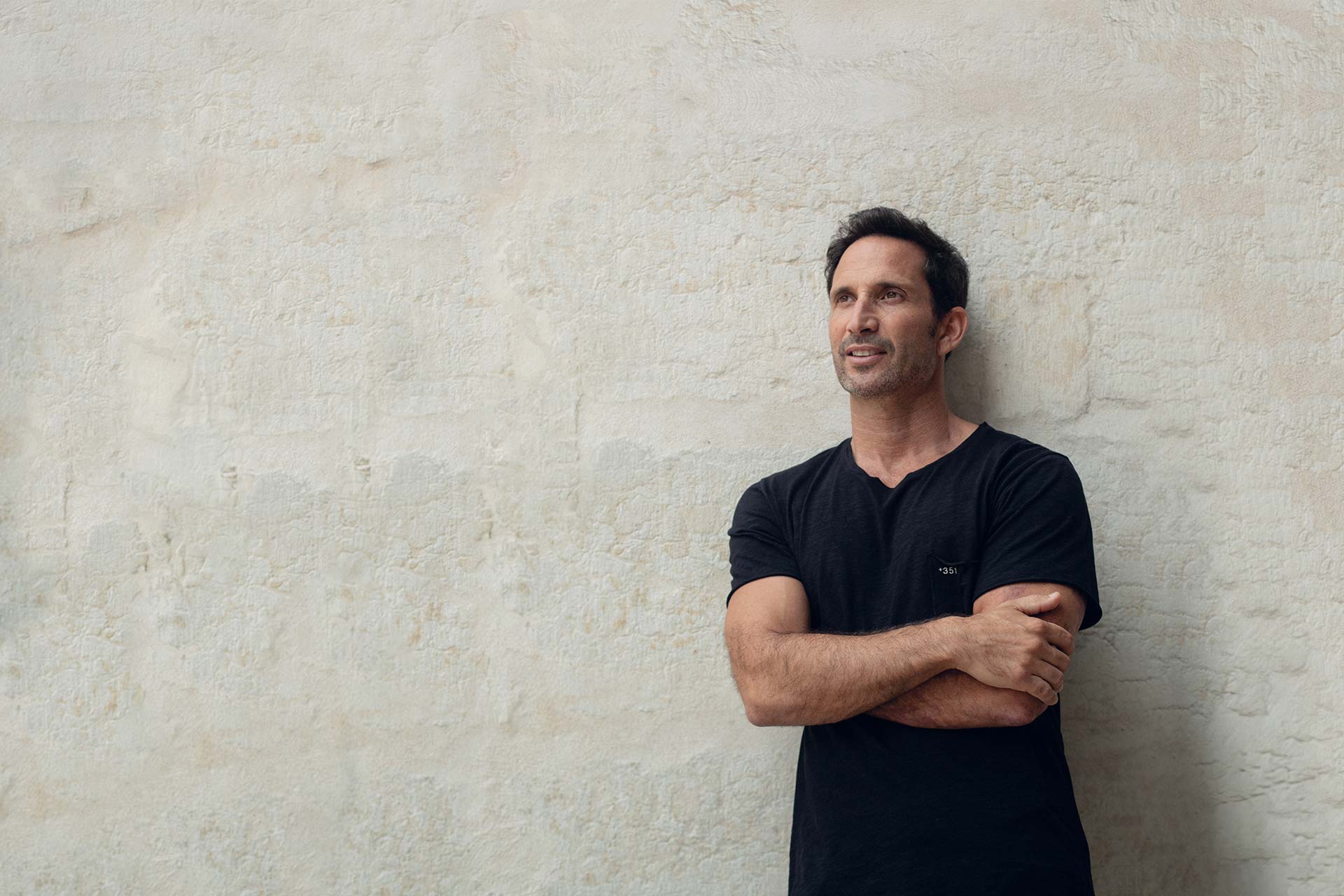 Man in a black t-shirt leans against a light stone wall with arms crossed, looking to the right.